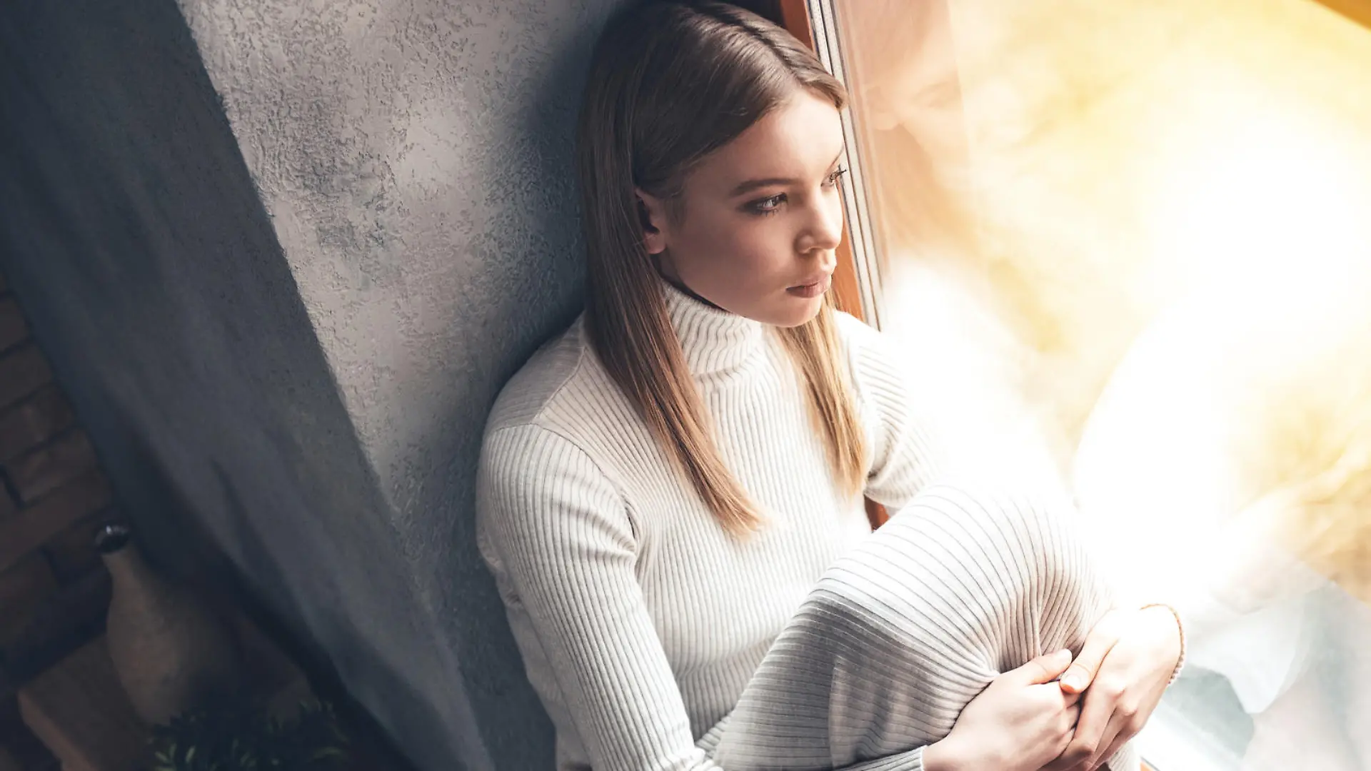 Top view of beautiful young woman looking through window while sitting at windowsill at home