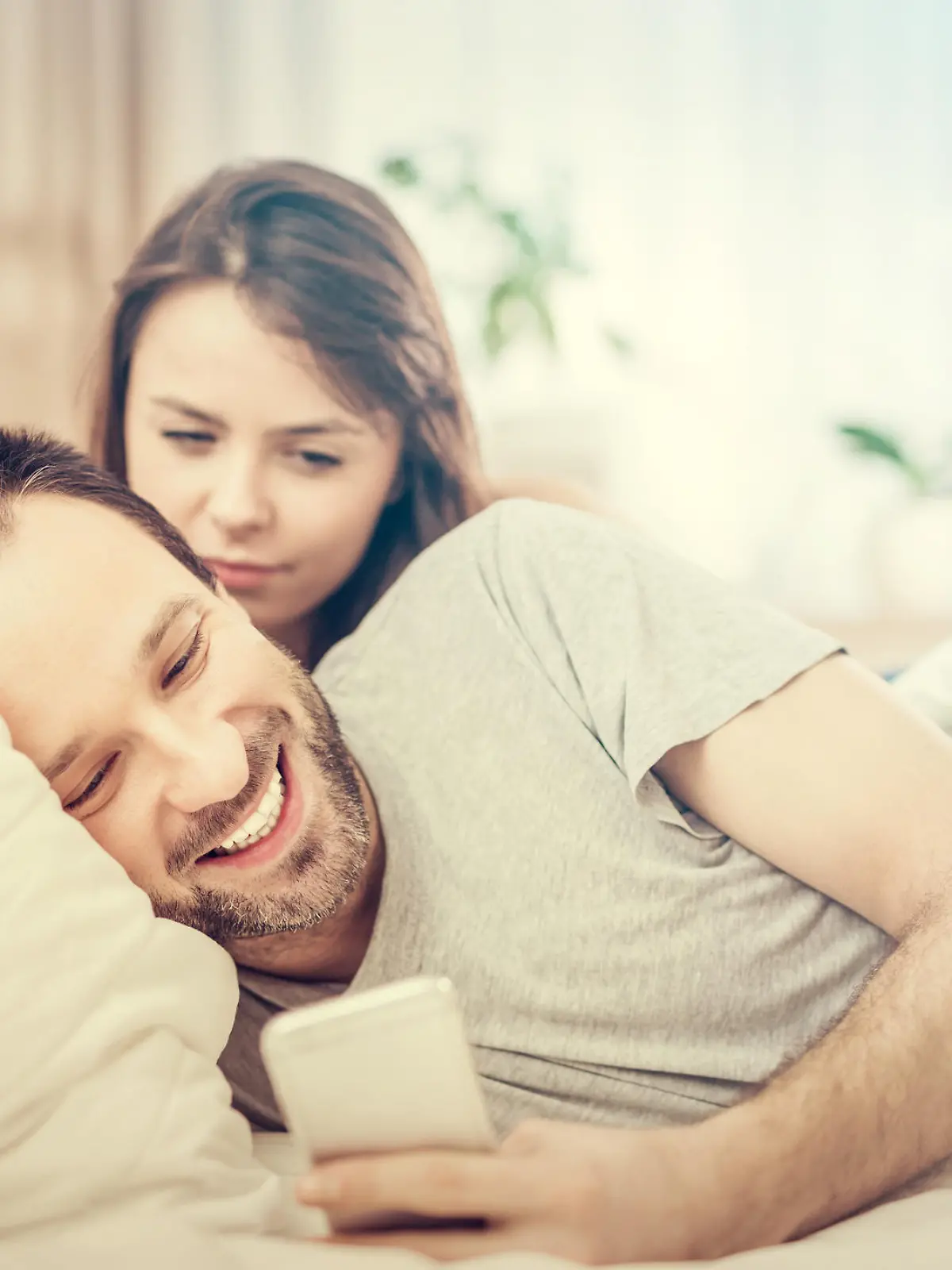 A young couple lying in bed. One of them use the phone and flirting. The other person is jealous and spies of the arm. The concept of jealousy and distrust.