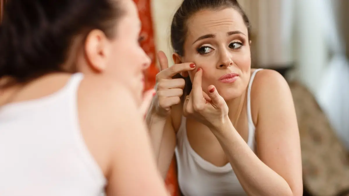 Unhappy young woman finding an acne on a cheek, sits before a mirror.