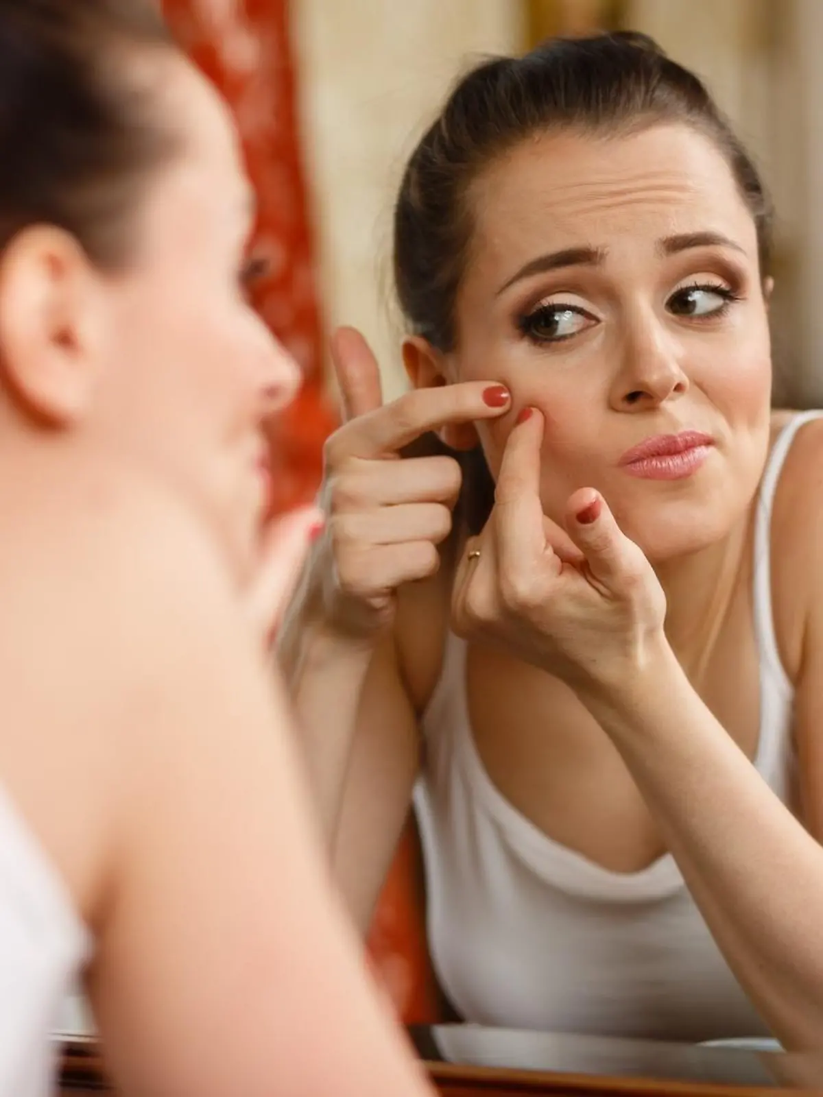 Unhappy young woman finding an acne on a cheek, sits before a mirror.