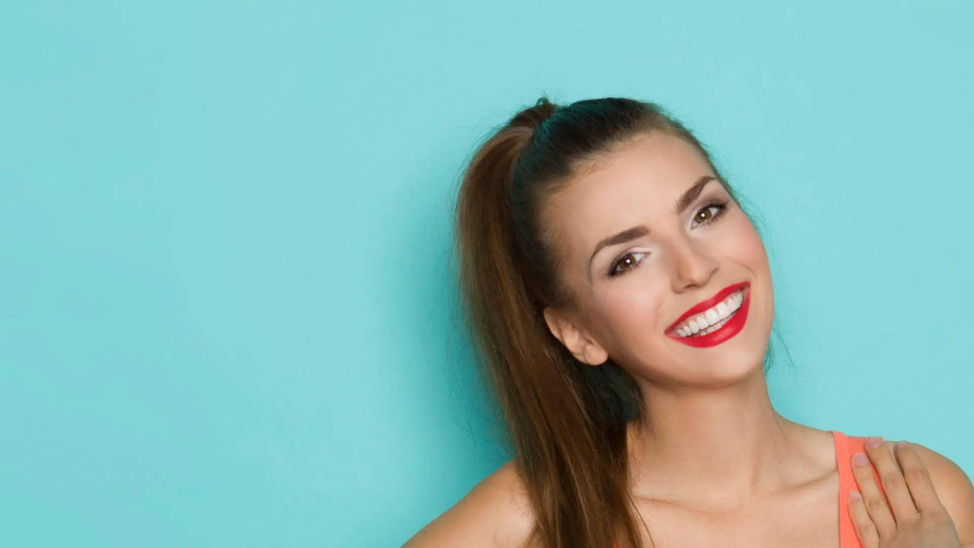 Sexy young woman in orange shirt with cleavage posing with hand on shoulder. Three quarter length studio shot on teal background.