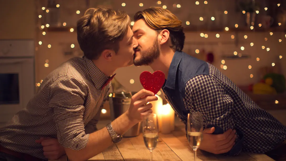 Loving gay couple having romantic date, drinking champagne and holding heart shape for Valentin's day. Domestic kitchen. Evening scene lit by candle and string lights.