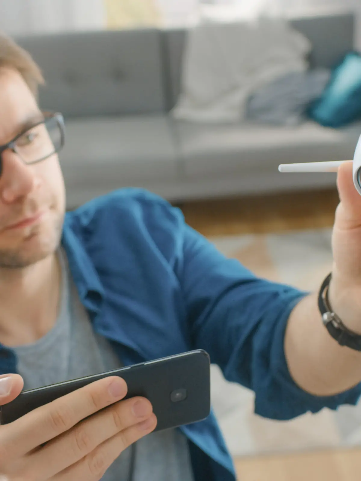 Young Man in Glasses Wearing a Blue Shirt is Adjusting a Modern Wi-Fi Surveillance Camera with Two Antennas on a White Wall at Home. He's Checking the Video Feed on his Smartphone.