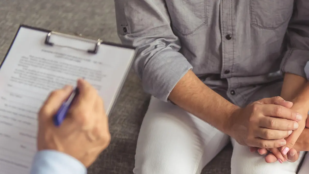 Cropped image of young couple holding hands while sitting on the couch at the psychotherapist. Doctor is making notes
