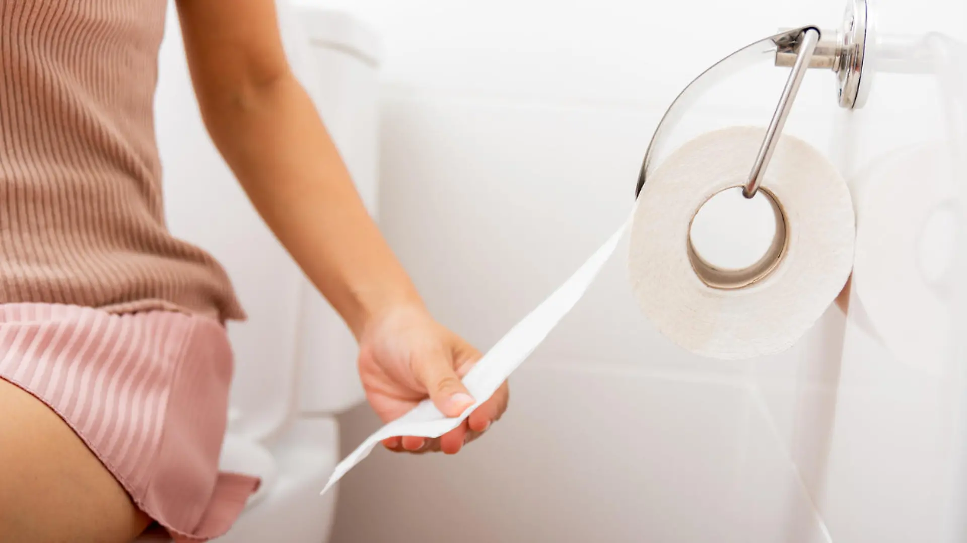 Closeup hand pulling toilet paper roll in holder for wipe, woman sitting on toilet she taking and tearing white tissue on wall to towel clean in bathroom, Healthcare concept