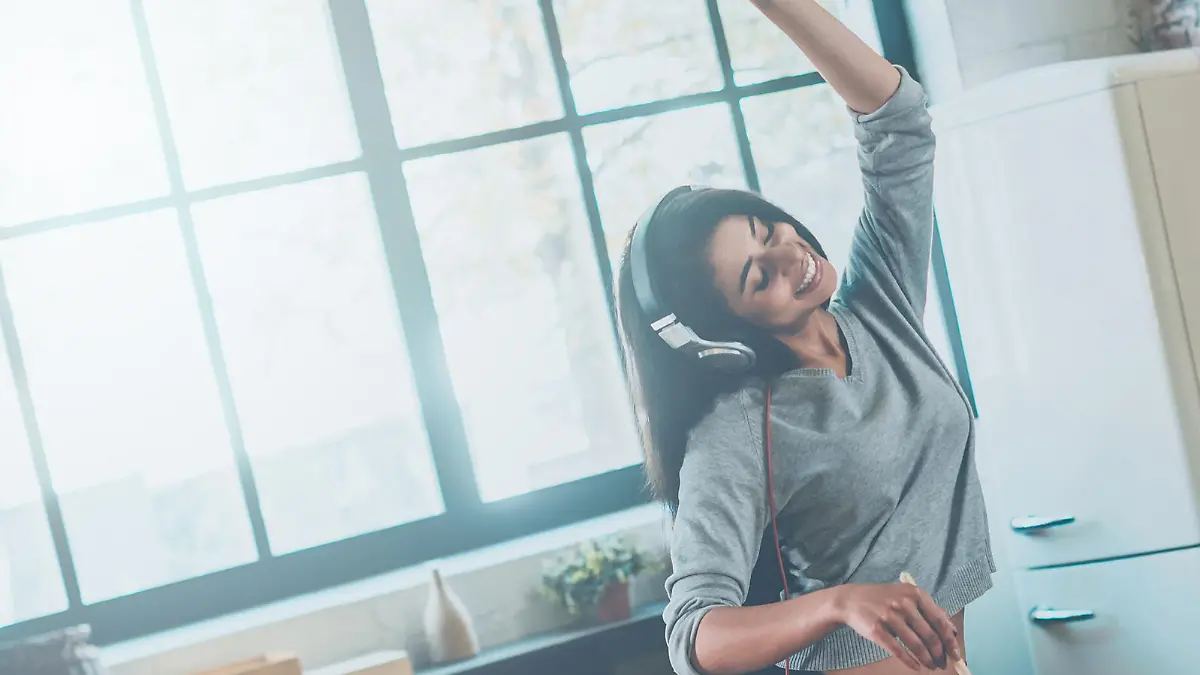 Beautiful young mixed race woman in headphones cooking salad and dancing while standing in kitchen at home