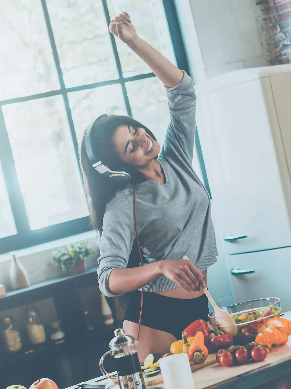 Beautiful young mixed race woman in headphones cooking salad and dancing while standing in kitchen at home