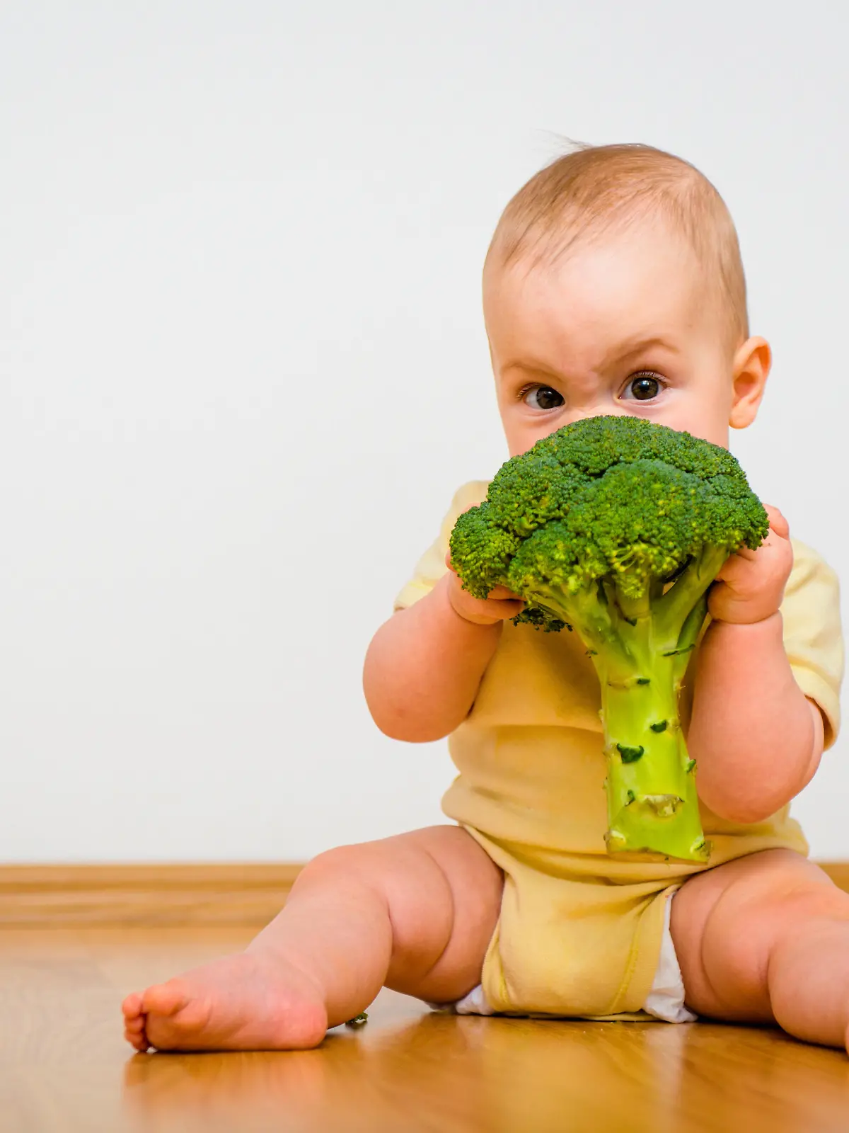 Cute baby eating broccoli sitting on floor indoors