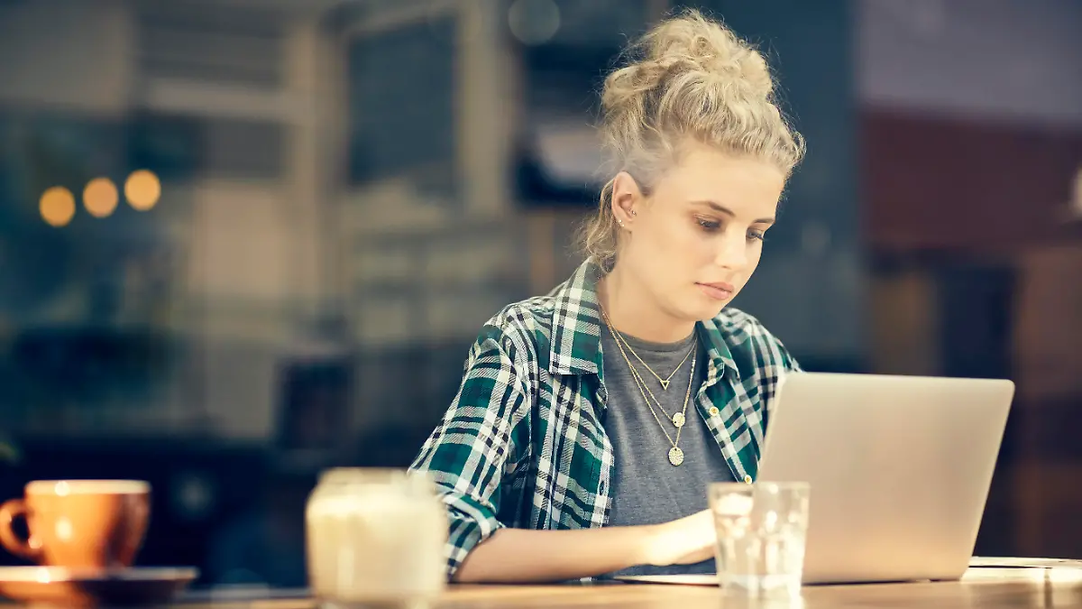 A photo of beautiful young woman using laptop at table inside cafe window. Female customer sitting in coffee shop. She is in trendy casuals.
