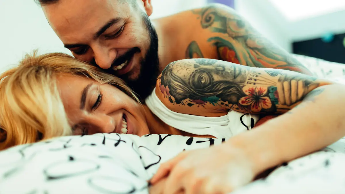 Young Tattooed Couple Sleeping together in their bedroom. They are cuddling and smiling. IKEA generic bed sheets.