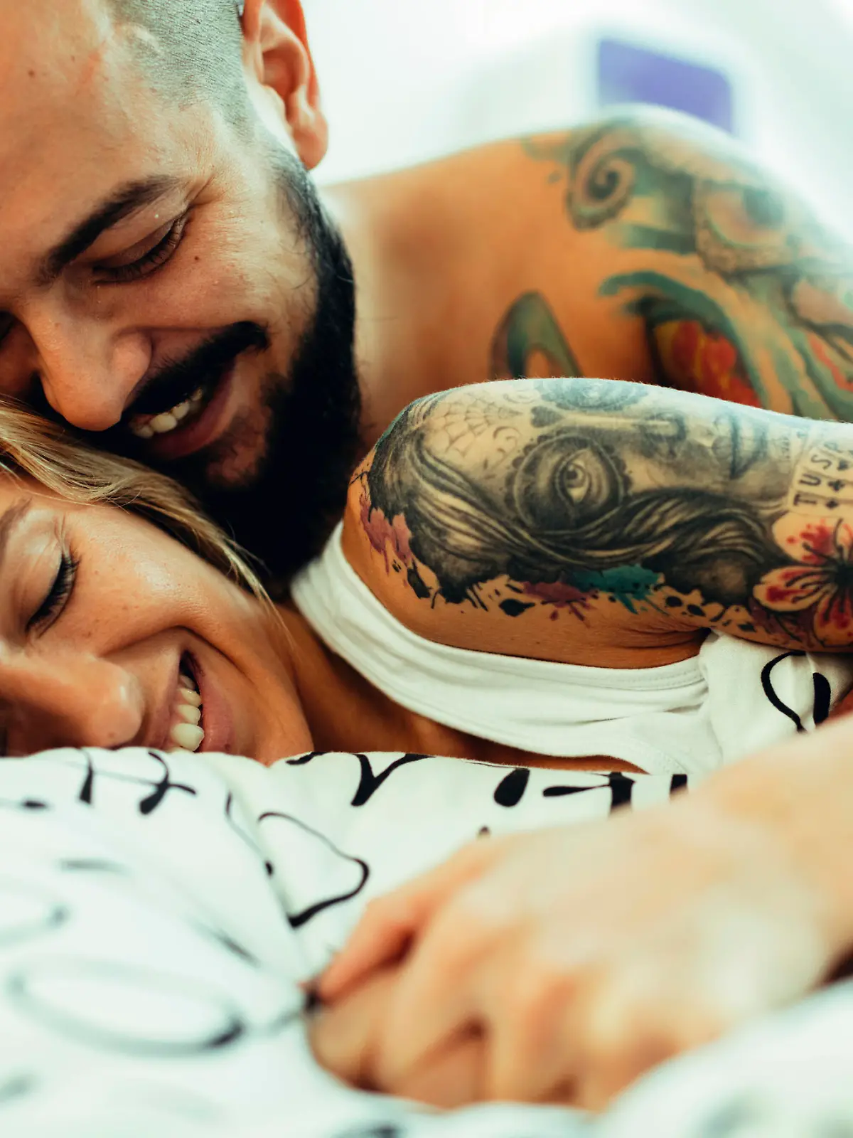 Young Tattooed Couple Sleeping together in their bedroom. They are cuddling and smiling. IKEA generic bed sheets.