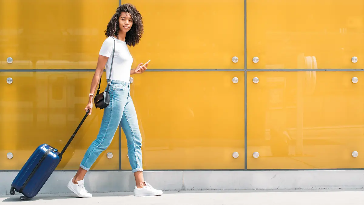 Woman using mobile phone while walking and pushing a suitcase in a yellow glass background