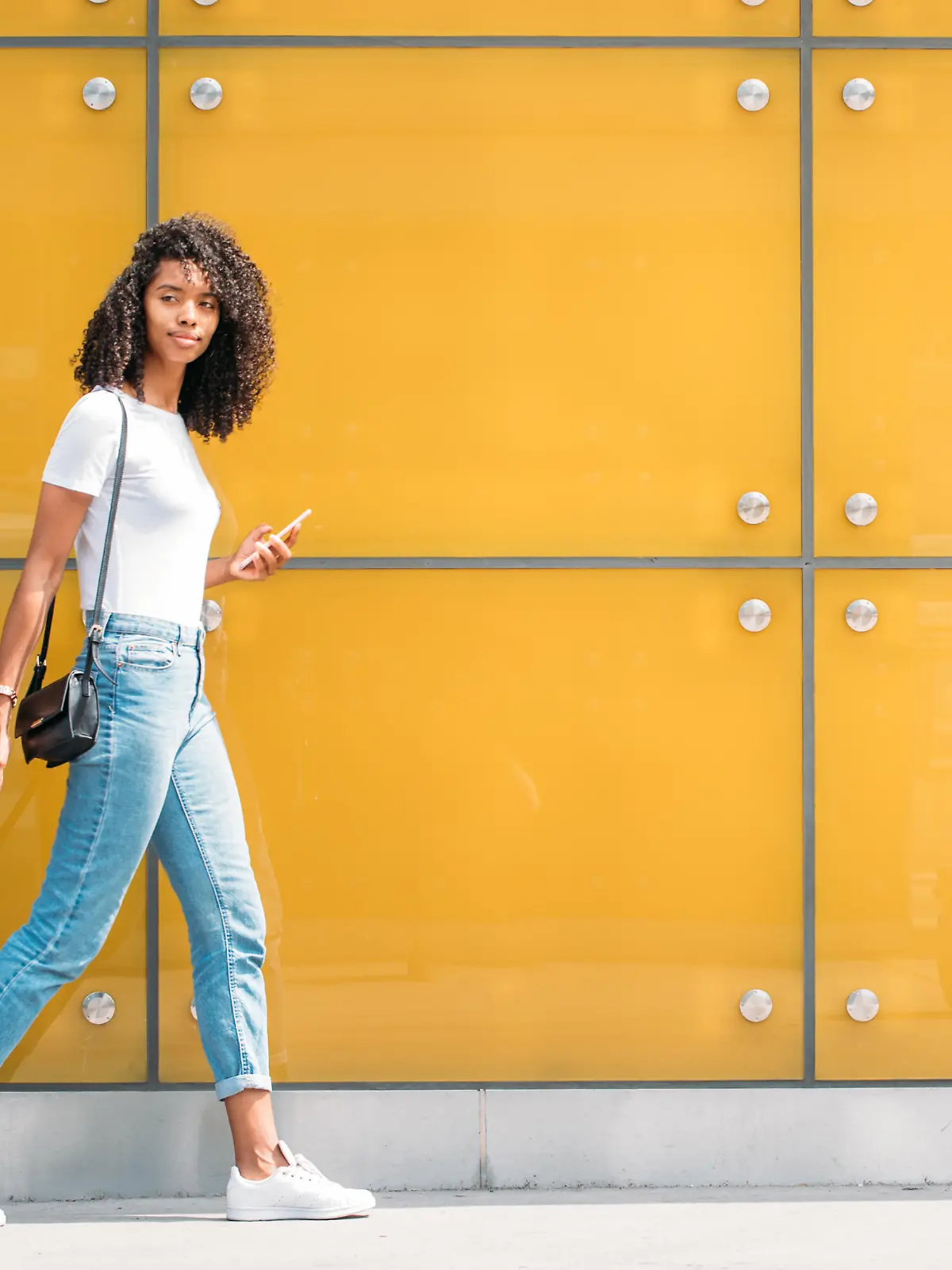 Woman using mobile phone while walking and pushing a suitcase in a yellow glass background