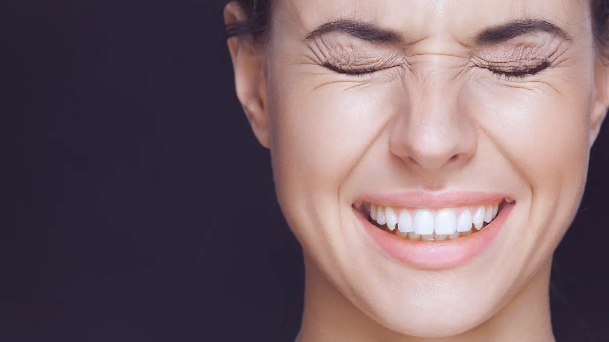 A close-up beauty portrait of a smiling young woman with strongly closed eyes. There are visible wrinkles around her eyes and mouth. The woman has healthy, clean skin, natural make-up and dark hair that is swept back from her face. The woman's shoulders are shirtless and visible, and she is isolated on a dark background. Developed from RAW; retouched with special care and attention; small amount of grain added for best final impression; ready made for print and web use;