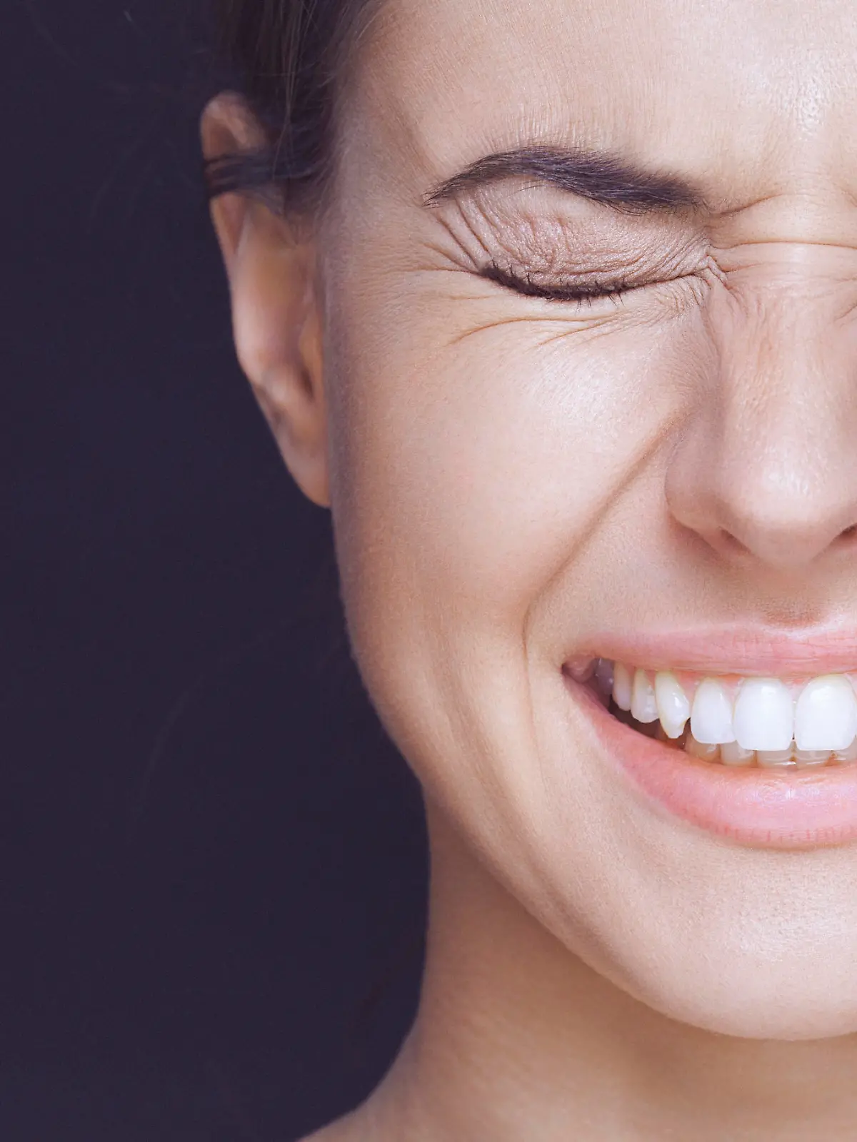 A close-up beauty portrait of a smiling young woman with strongly closed eyes. There are visible wrinkles around her eyes and mouth. The woman has healthy, clean skin, natural make-up and dark hair that is swept back from her face. The woman's shoulders are shirtless and visible, and she is isolated on a dark background. Developed from RAW; retouched with special care and attention; small amount of grain added for best final impression; ready made for print and web use;