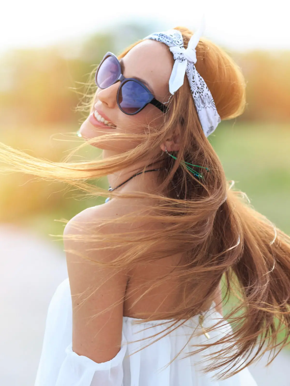 Young smilling woman posing on the road. Wears white blouse, headband and sunglasses, flowing hair. Focus on foreground.