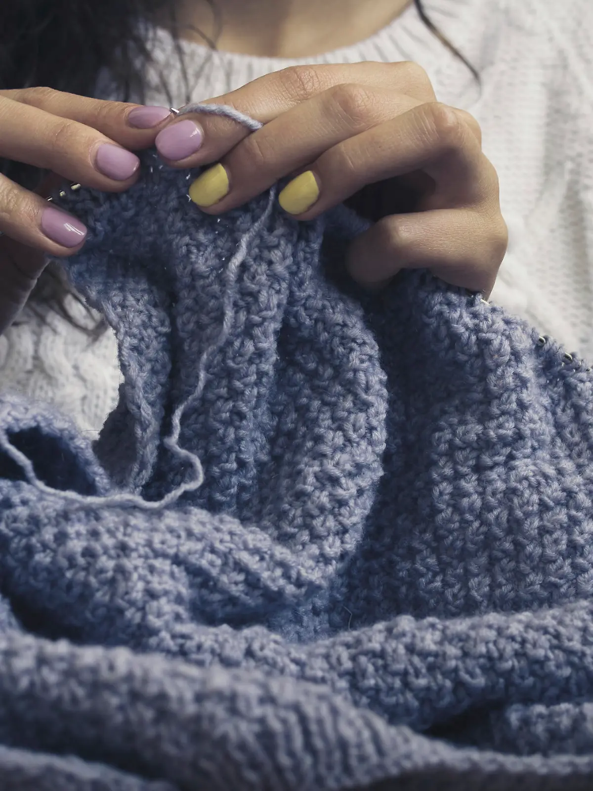 The girl in the white sweater with bright manicure knits blue thread. Vintage photo close up.