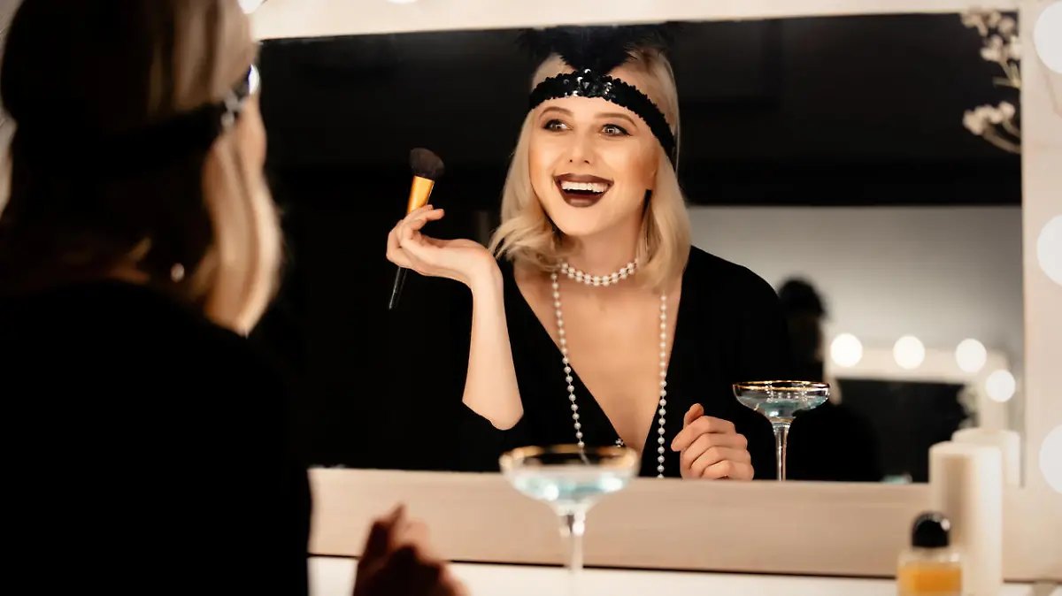 Beautiful blonde woman in twenties years clothes applying makeup near a mirror with bulbs