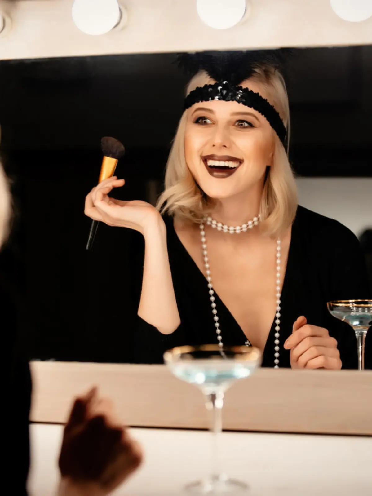 Beautiful blonde woman in twenties years clothes applying makeup near a mirror with bulbs