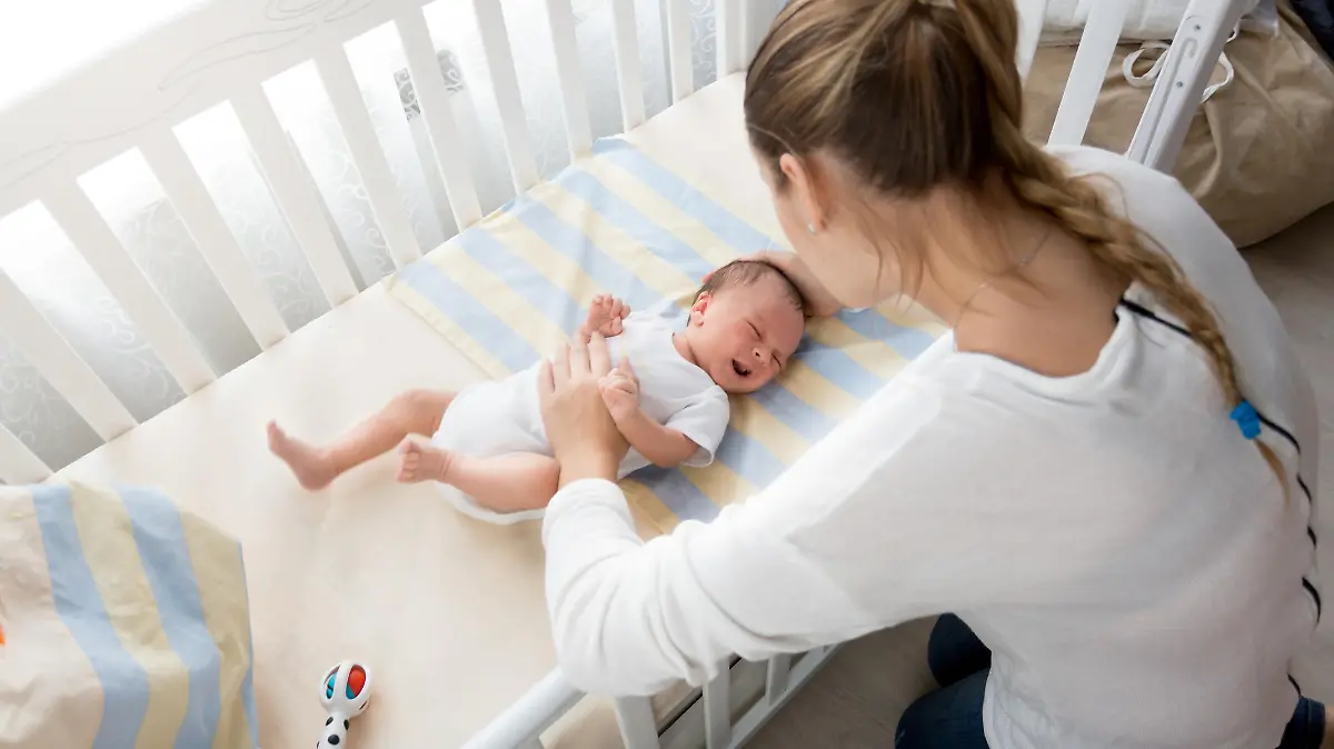 Young mother sitting near the cradle and holding baby's hand