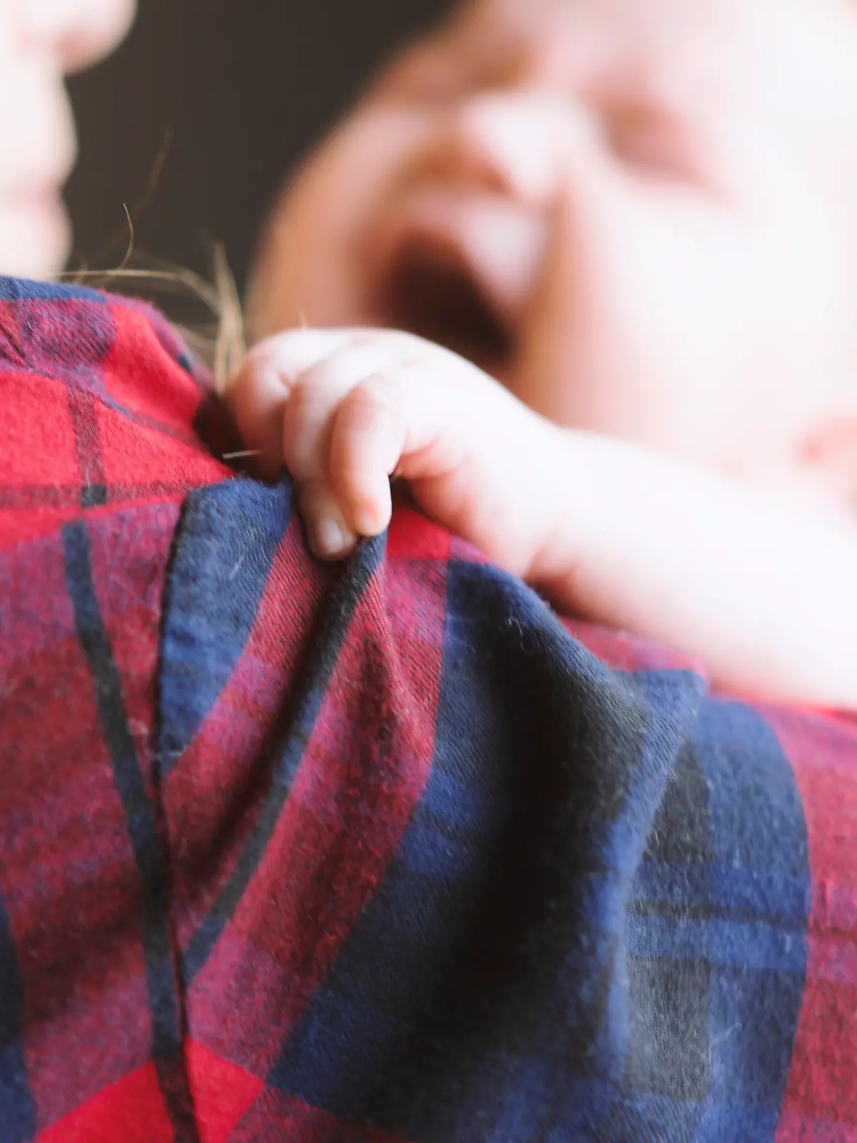 Newborn baby wearing red and white striped bodysuit, in her mother's arms, crying. Mother wearing red and blue plaid shirt and blue jeans, holding baby with care. Baby holding mothers shirt, focus on baby's hand.