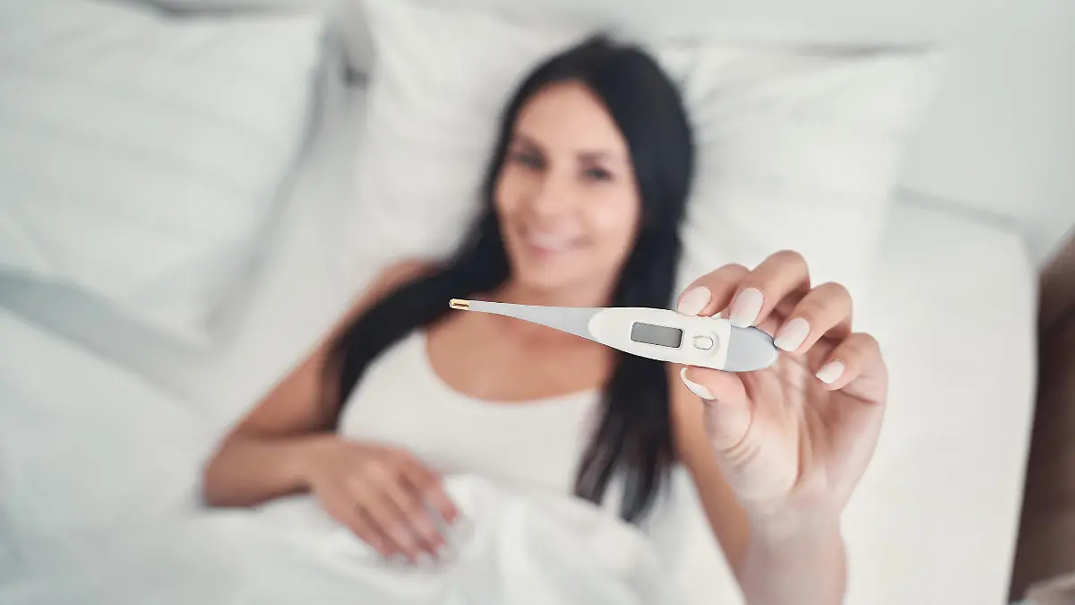 Basaltemperatur messen: Frau liegt im Bett und hält Thermometer in der Hand.