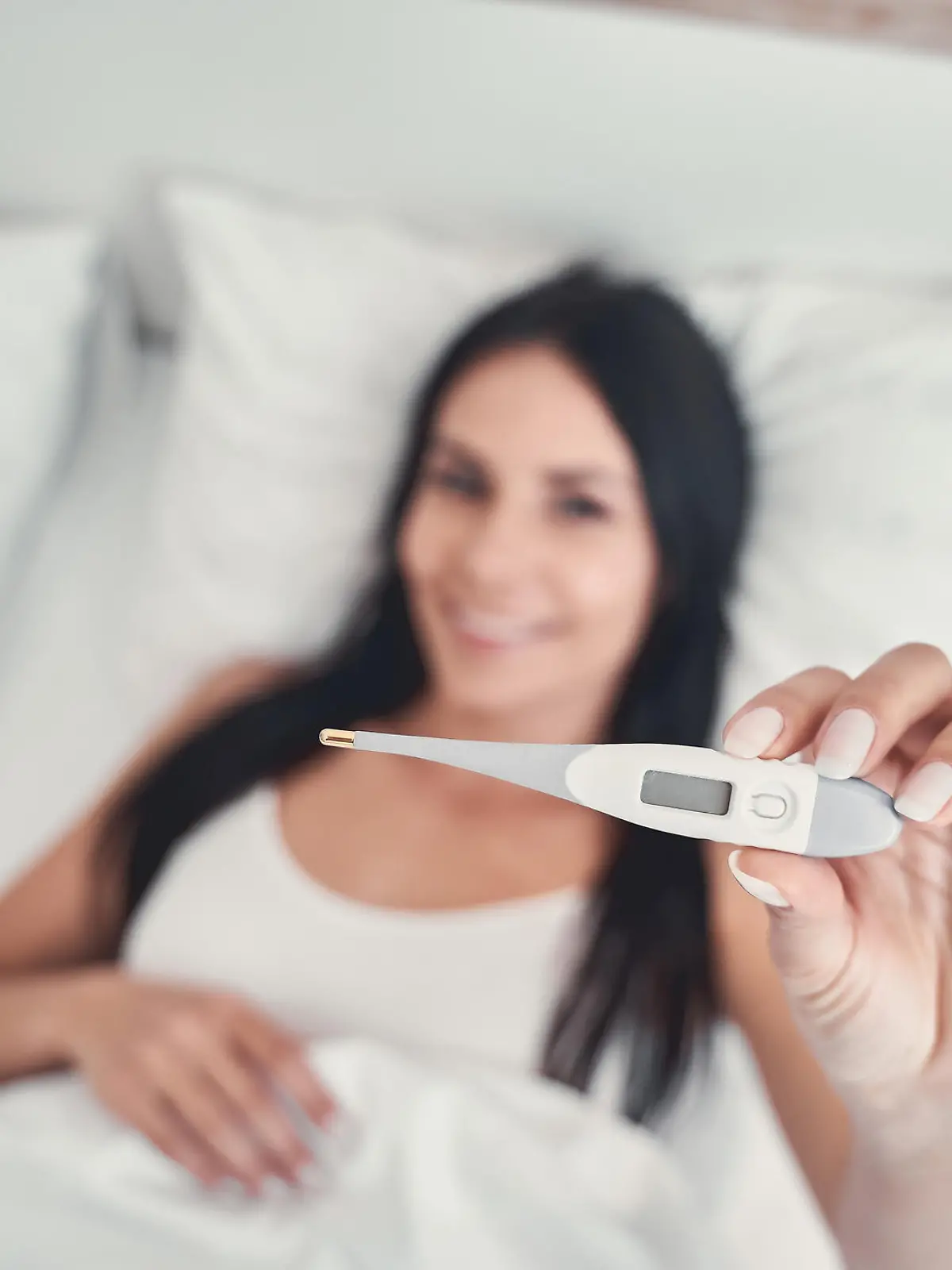 Basaltemperatur messen: Frau liegt im Bett und hält Thermometer in der Hand.