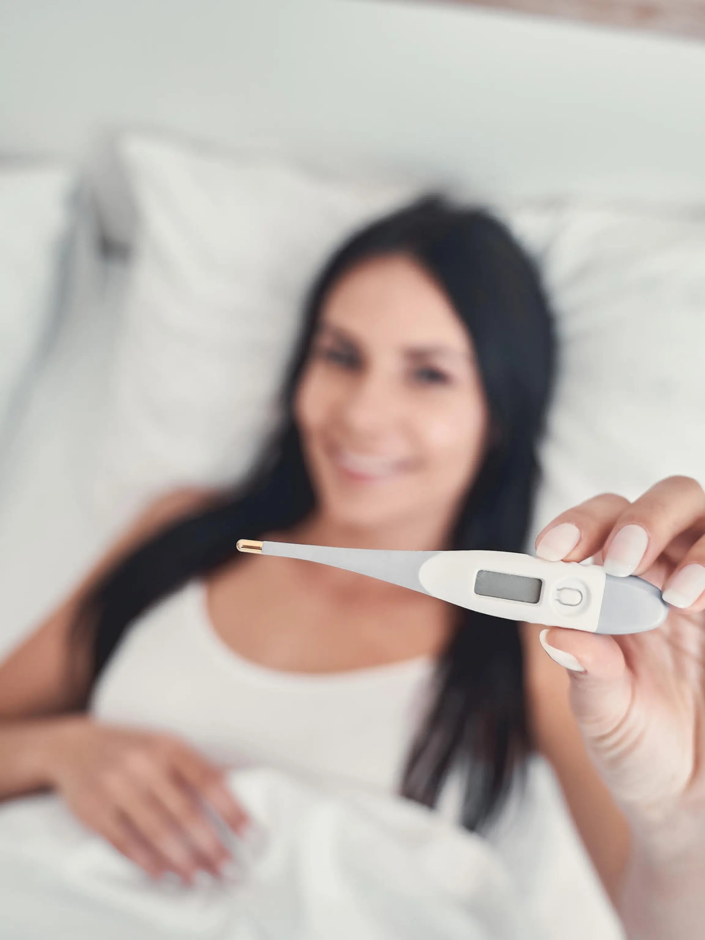Basaltemperatur messen: Frau liegt im Bett und hält Thermometer in der Hand.