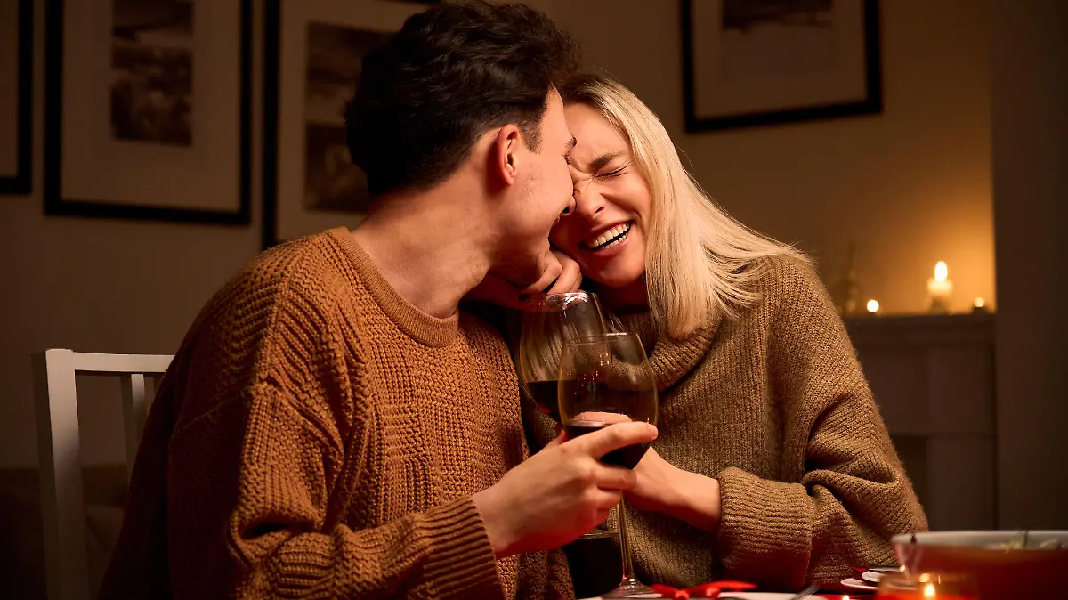 Happy young couple in love hugging, laughing, drinking wine, enjoying talking, having fun together celebrating Valentines day dining at home, having romantic dinner date with candles sitting at table.