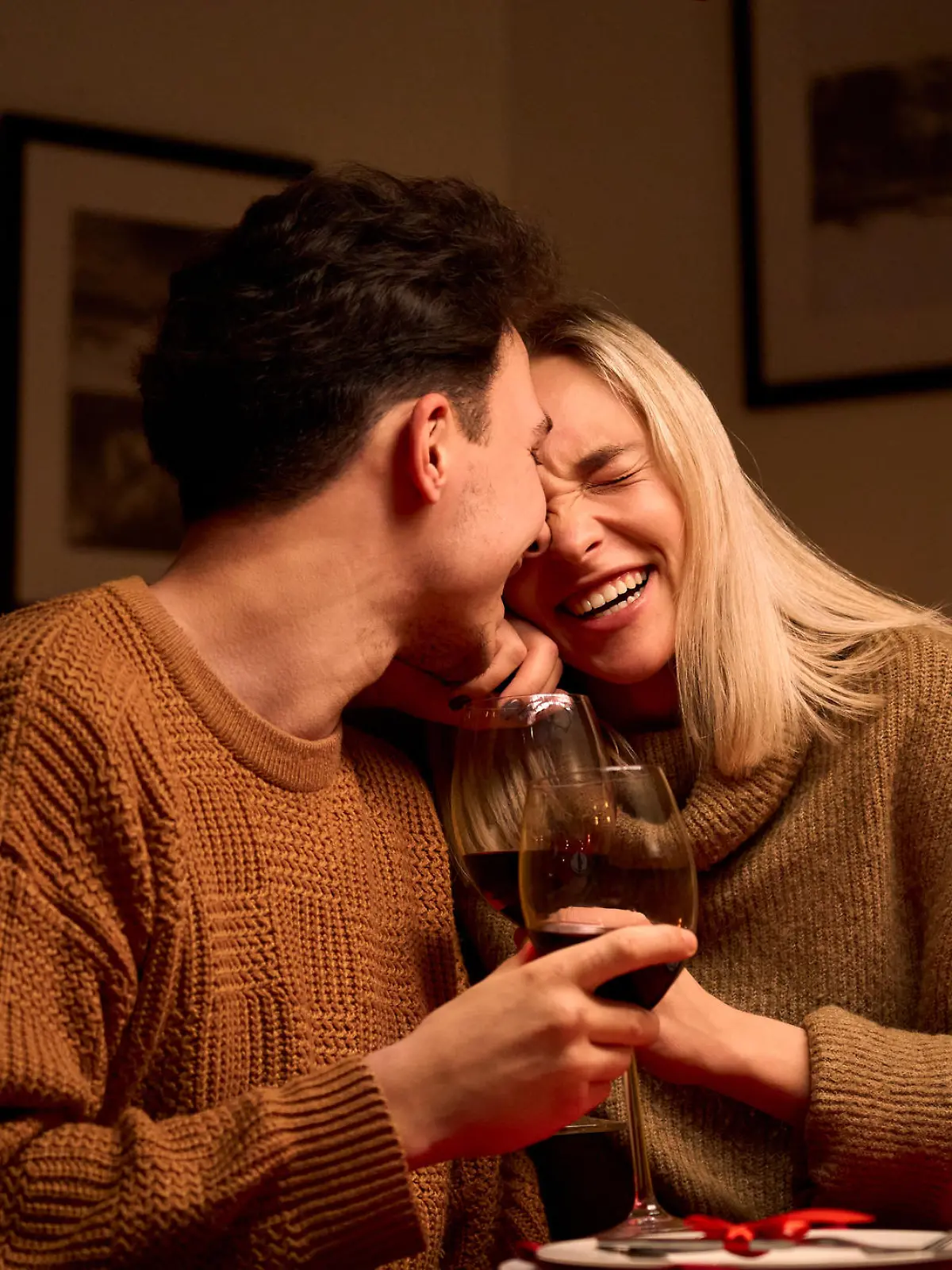 Happy young couple in love hugging, laughing, drinking wine, enjoying talking, having fun together celebrating Valentines day dining at home, having romantic dinner date with candles sitting at table.
