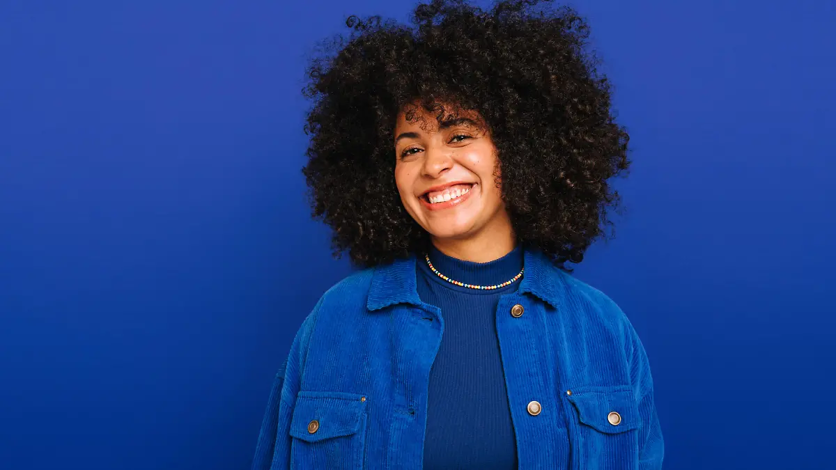 Feeling good in blue. Happy woman with curly hair smiling at the camera while standing against a blue background. Beautiful woman with curly hair wearing her natural air with pride.