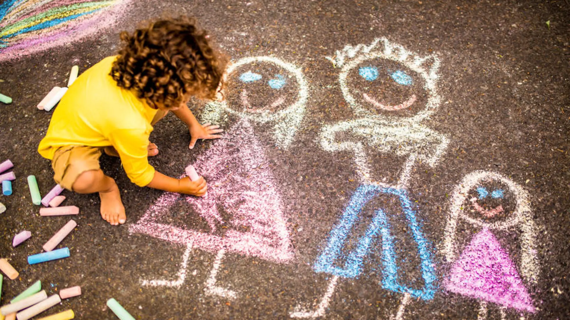 Overhead shot of a little boy drawing a  picture of his family in colourful chalk on tarmac