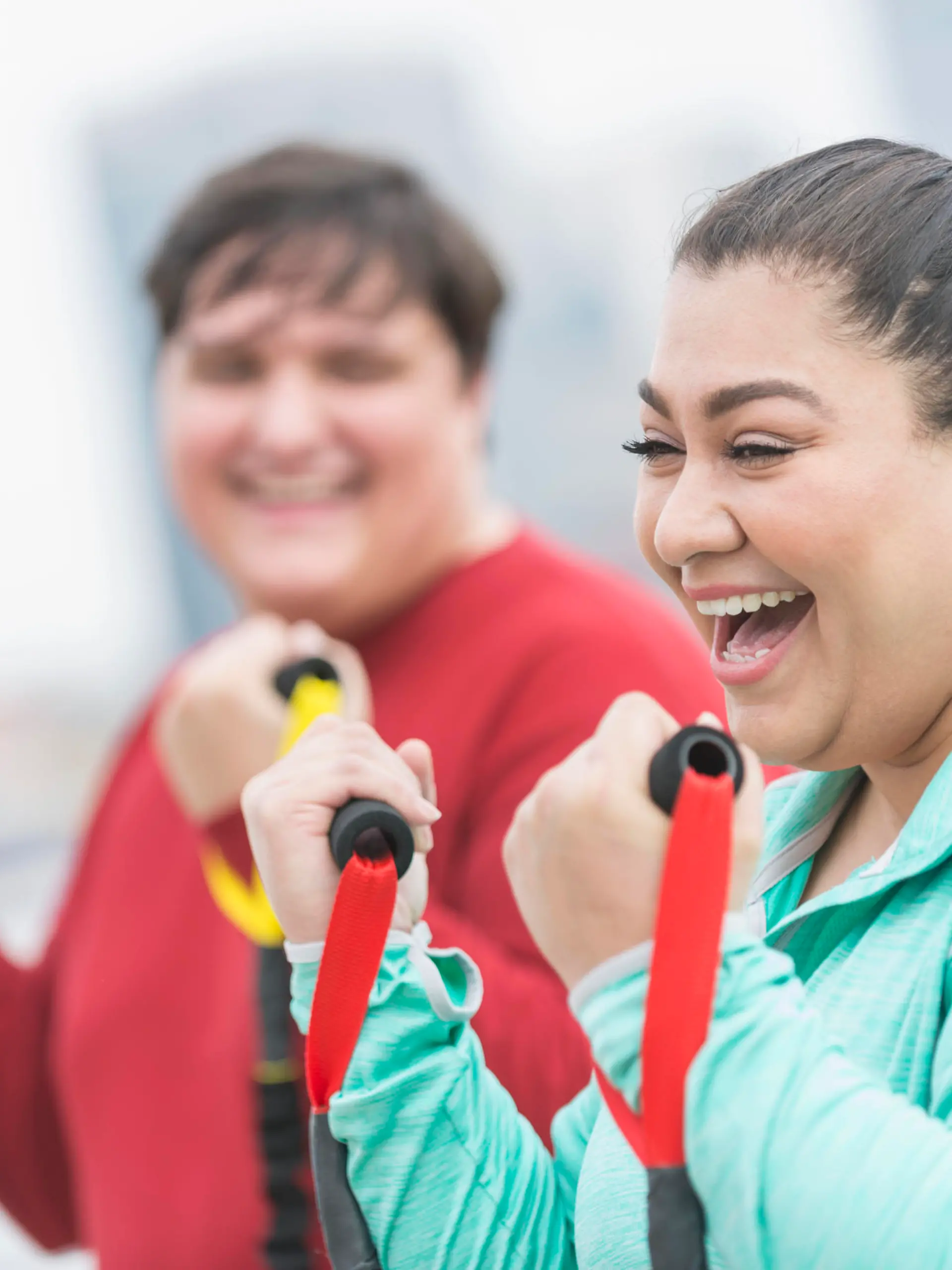 An overweight Hispanic woman in her 30s exercising outdoors with resistance bands. Her friend is out of focus in the background. She is laughing, having fun.