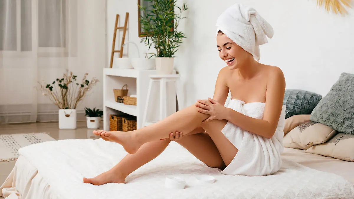 Young beautiful woman in towel applying cream on her legs after shower at home