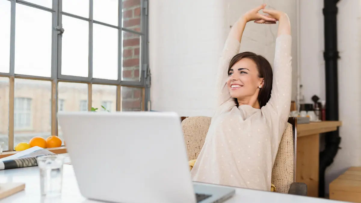 Happy relaxed young woman sitting in her kitchen with a laptop in front of her stretching her arms above her head and looking out of the window with a smile
