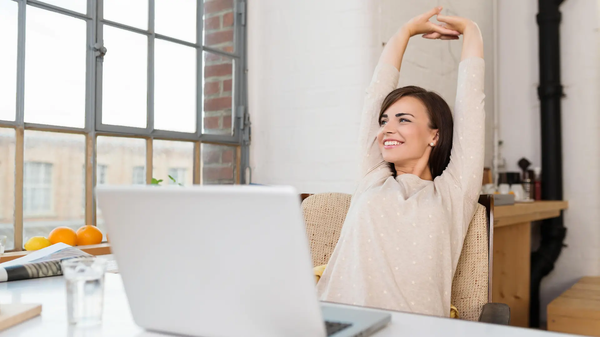 Happy relaxed young woman sitting in her kitchen with a laptop in front of her stretching her arms above her head and looking out of the window with a smile