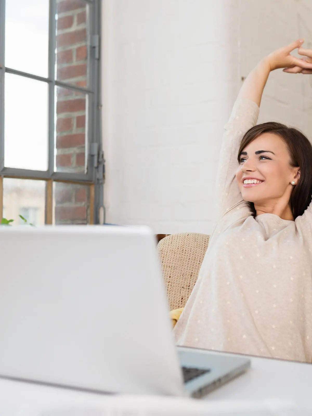 Happy relaxed young woman sitting in her kitchen with a laptop in front of her stretching her arms above her head and looking out of the window with a smile