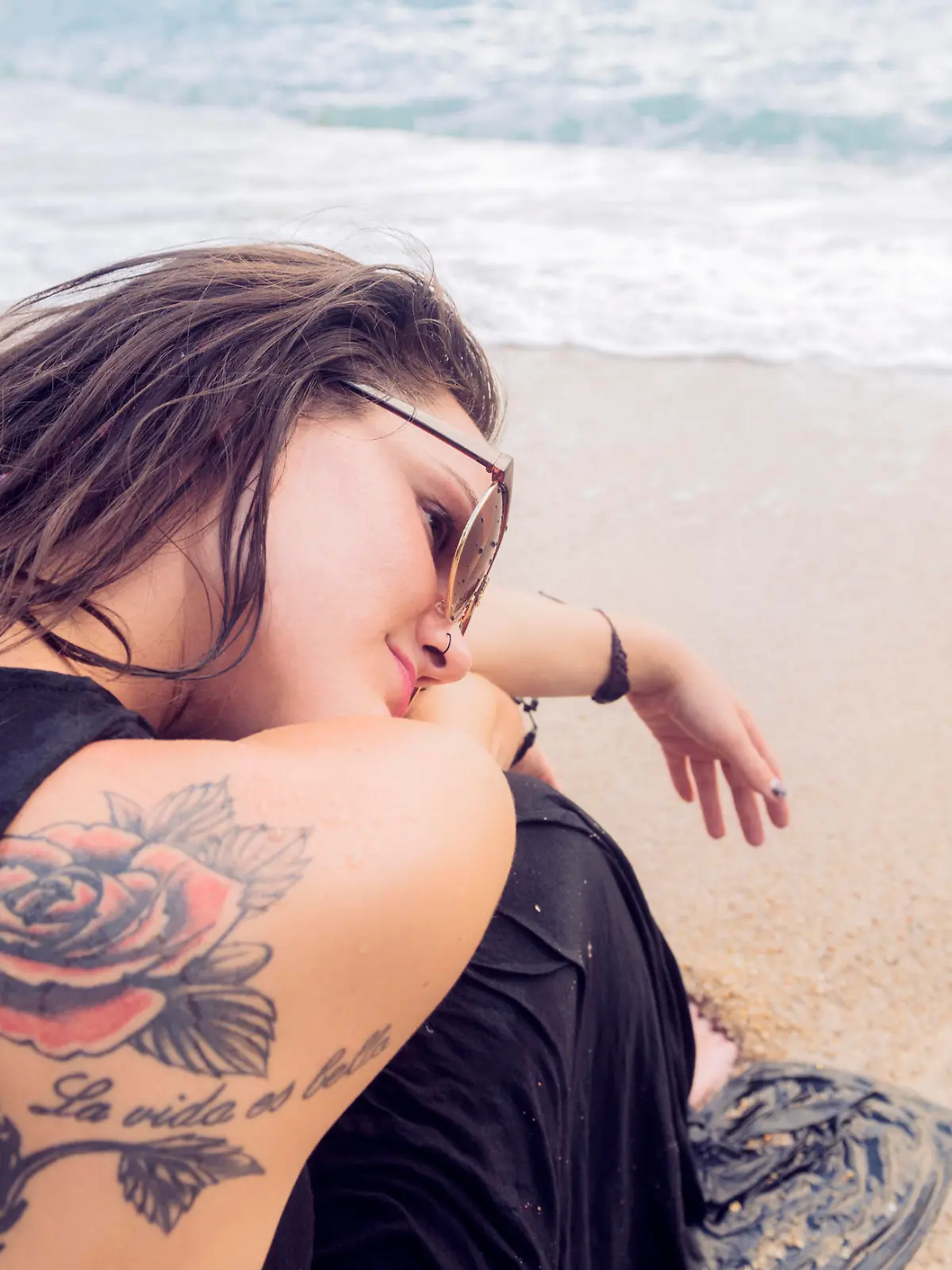 This royalty free, close up, color, stock photograph is of a beautiful Canadian woman in her mid 20s sitting on the sandy shore of a tropical beach on Koh Phangan, Thailand. She wears a black dress and rests her head on top of her arms with her knees bent into her chest. A tattoo of a rose is visible on the back of her shoulder. Photographed from a rear high angle view with a Nikon D800 DSLR camera.