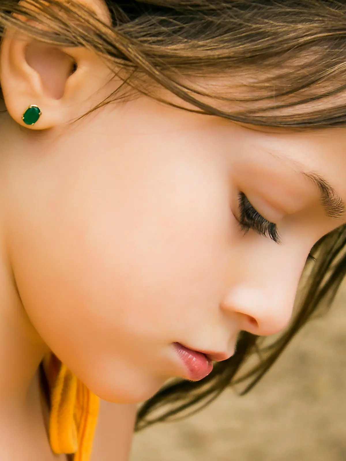 A delicate DSLR close-up photo of a cute 6-year-old caucasian girl. She is in profile to the camera looking down at something out of the frame. She is wearing a green earring and a yellow spaghetti straps blouse. Her soft skin shines.