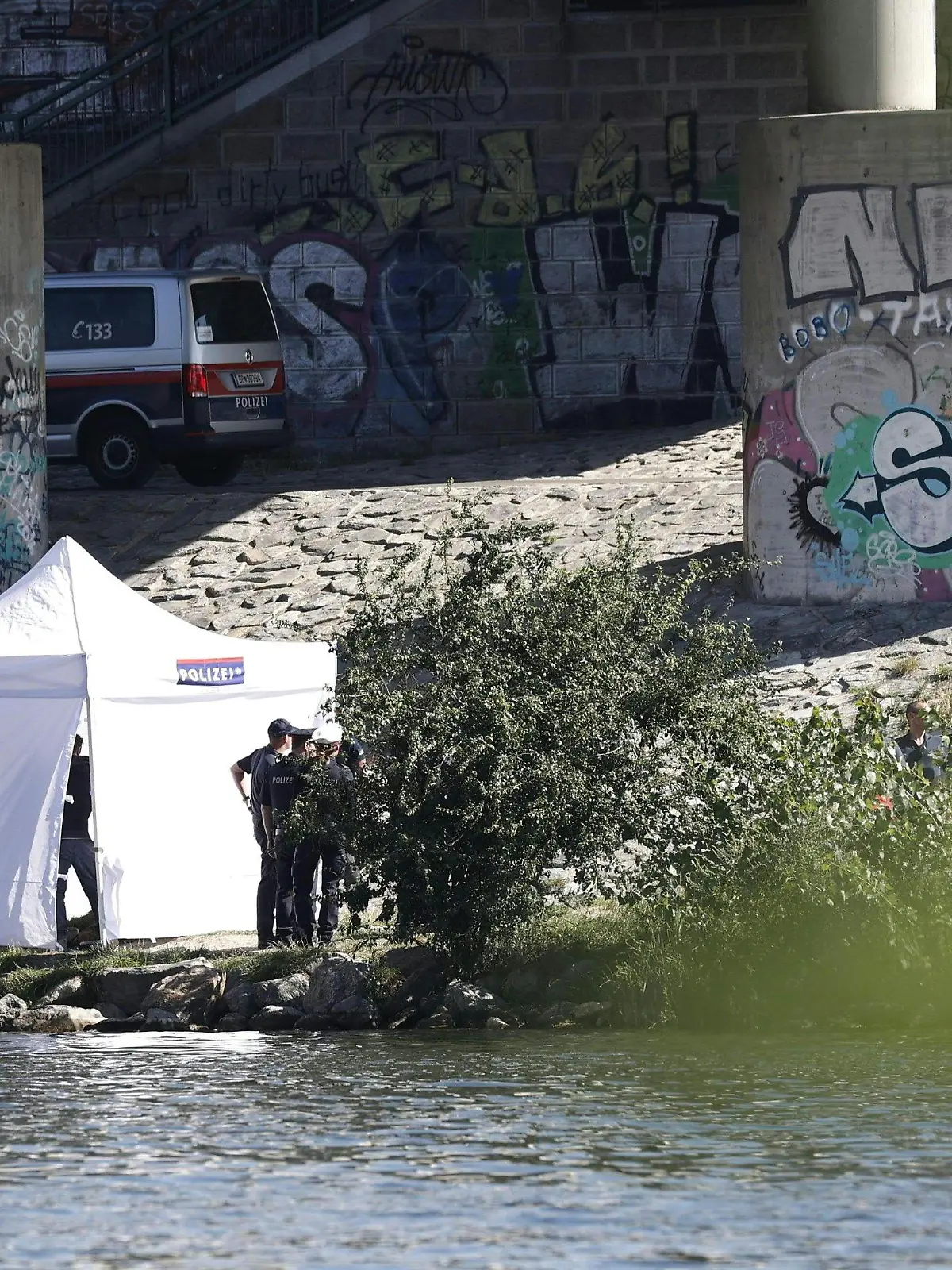 14.08.2023, Österreich, Wien: Einsatzkräfte ermitteln am Flussufer der Neuen Donau. Bei einer Hammerattacke wurde eine Frau verletzt - ihr vermisster fünfjähriger Sohn später tot in einem Seitenarm der Donau gefunden. Am Montag wurde dort eine weitere Leiche entdeckt. Foto: Tobias Steinmaurer/APA/dpa +++ dpa-Bildfunk +++