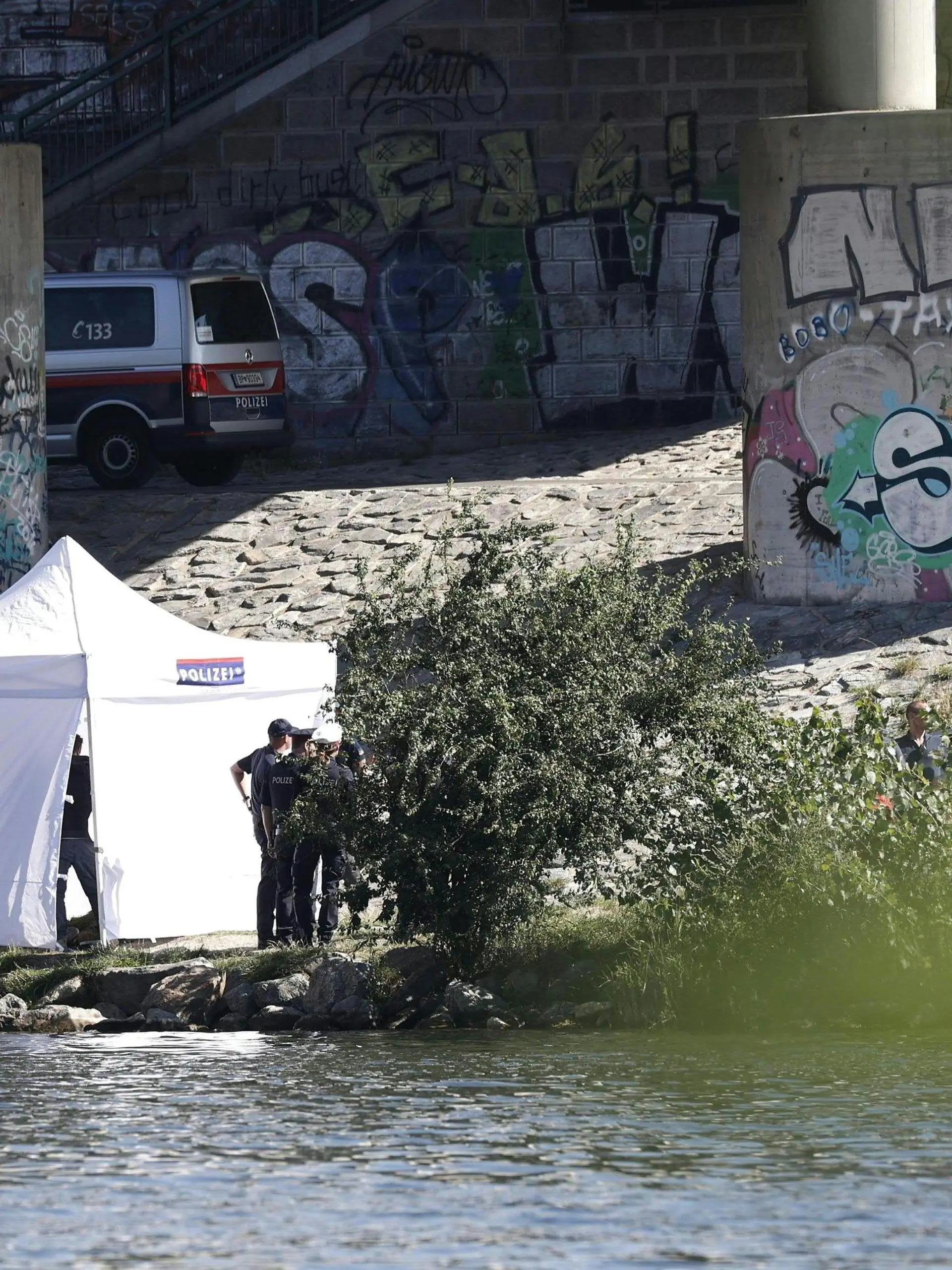 14.08.2023, Österreich, Wien: Einsatzkräfte ermitteln am Flussufer der Neuen Donau. Bei einer Hammerattacke wurde eine Frau verletzt - ihr vermisster fünfjähriger Sohn später tot in einem Seitenarm der Donau gefunden. Am Montag wurde dort eine weitere Leiche entdeckt. Foto: Tobias Steinmaurer/APA/dpa +++ dpa-Bildfunk +++