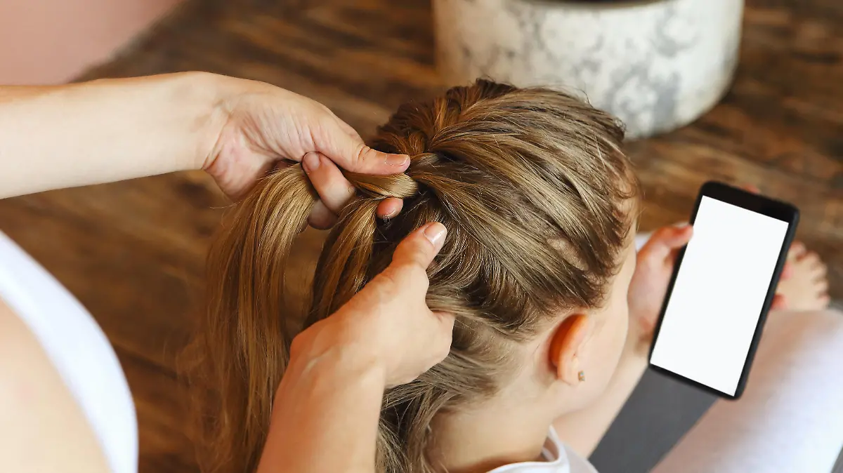 Young blonde girl getting her long hair braided by woman hairdresser before sport training while looking at cell phone with mockup screen and sitting on floor at home or fitness studio || Modellfreigabe vorhanden