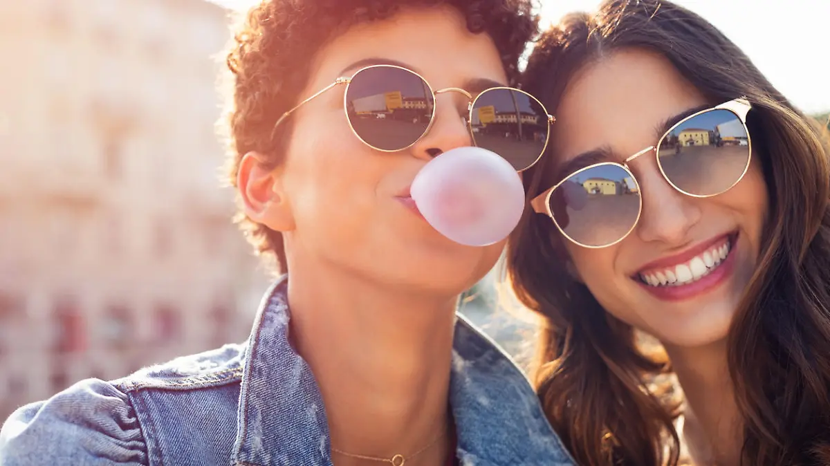Portrait of brazilian woman with curly hair making bubble while latin girl smiling. Multiethnic friends enjoying vacation in city and looking at camera. Lesbian couple having fun while inflating chewing gum and making pink balloon.