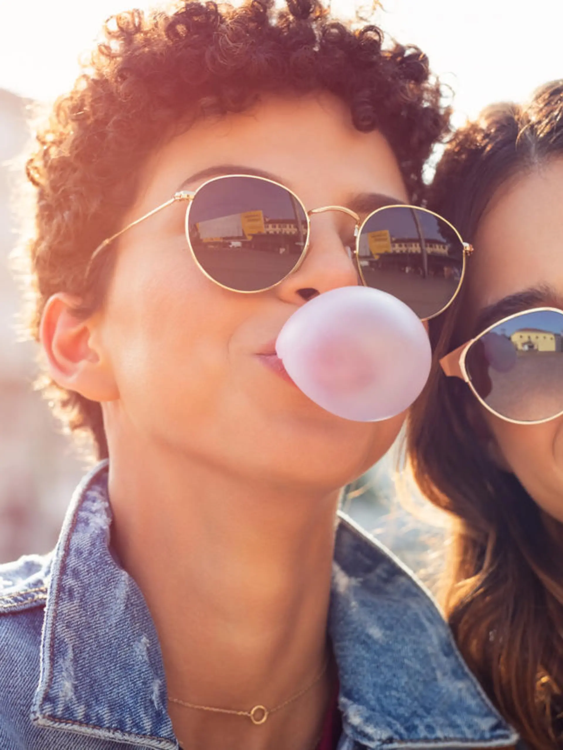 Portrait of brazilian woman with curly hair making bubble while latin girl smiling. Multiethnic friends enjoying vacation in city and looking at camera. Lesbian couple having fun while inflating chewing gum and making pink balloon.