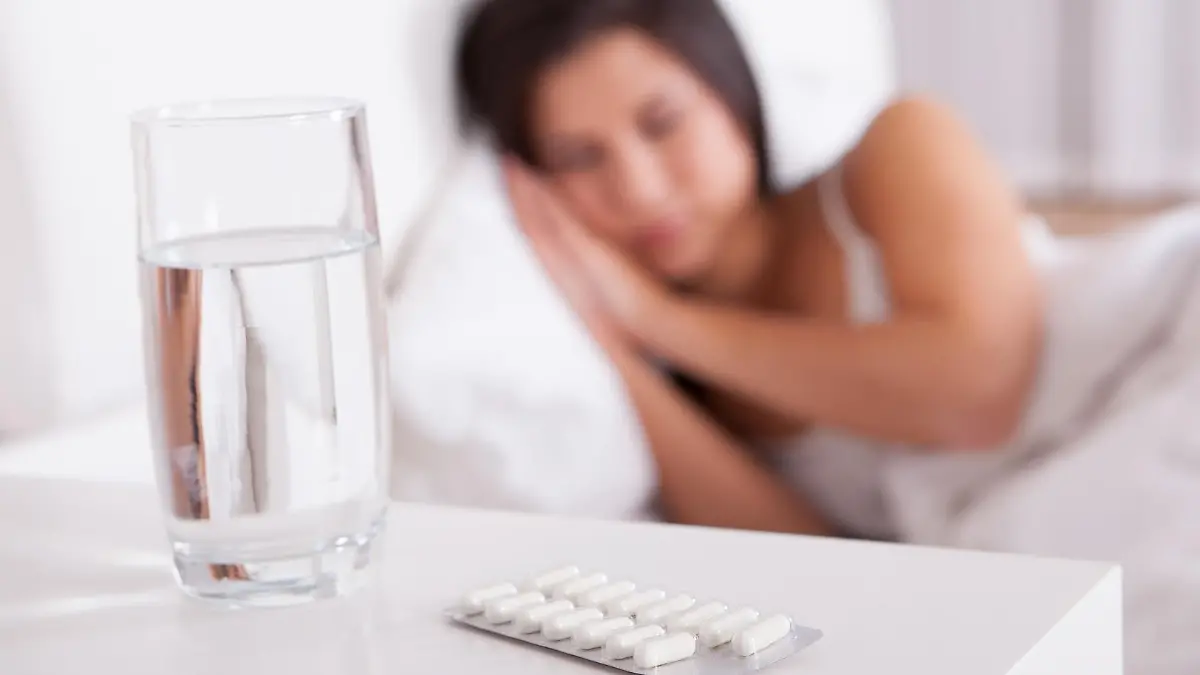 Sick woman resting in bed with focus to a glass of water and a blister pack of tablets and medication lying on the table alongside her bed
