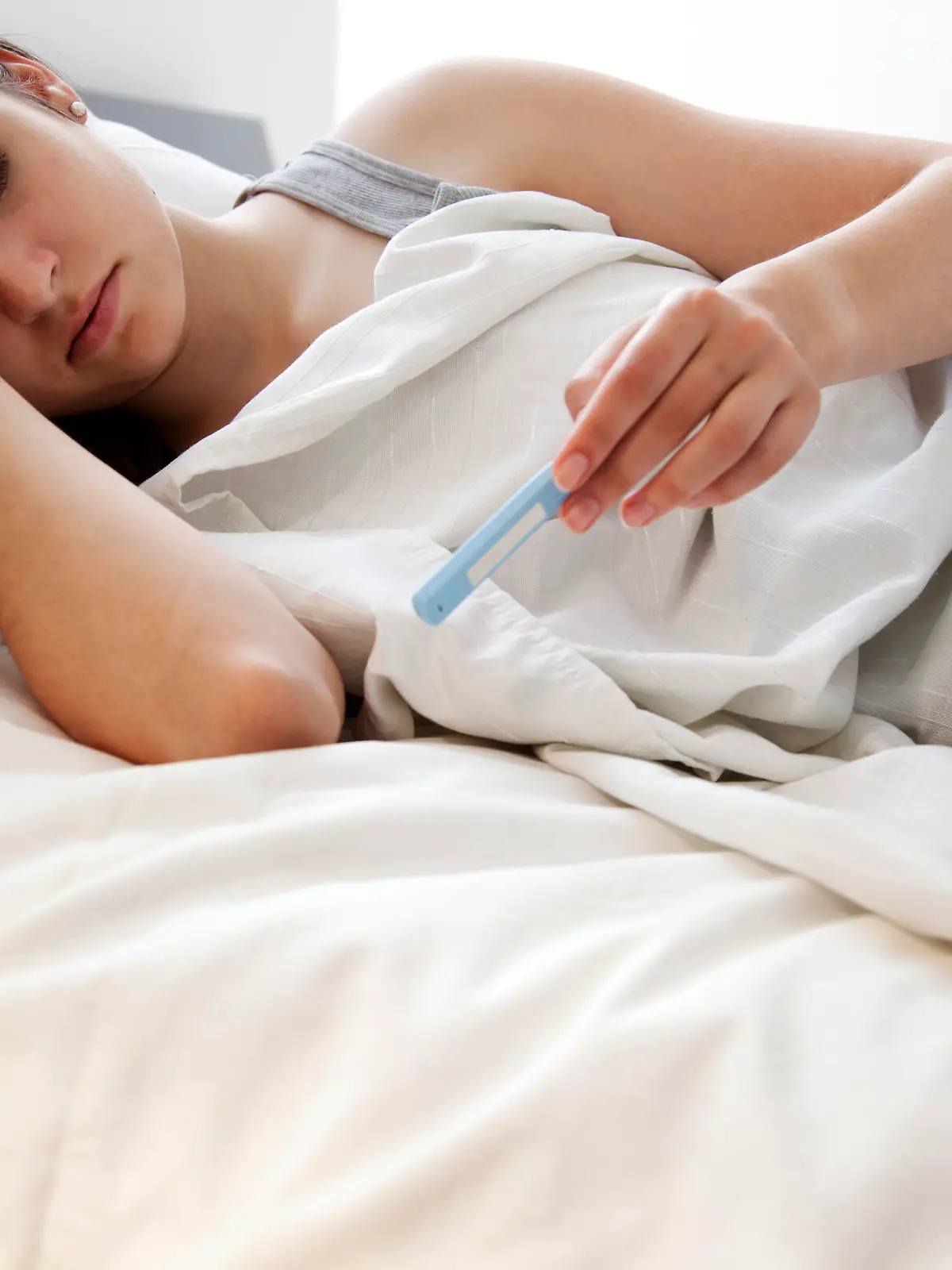 Eine Frau liegt krank, inklusive Fieber-Thermometer, im Bett.