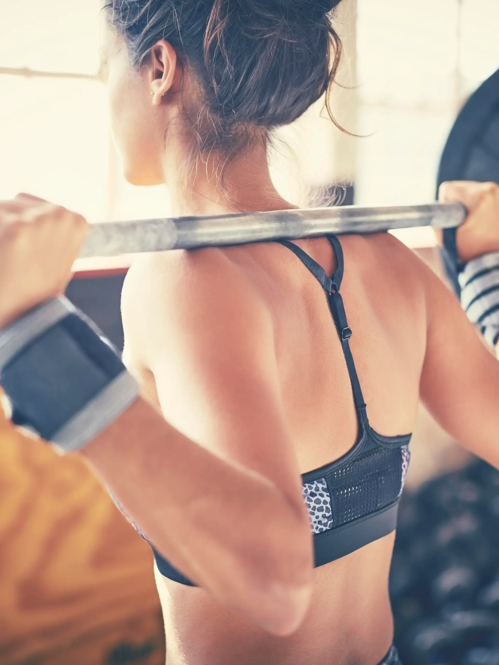 Rear view of woman exercising with barbell. Young female is lifting weights. She is standing in brightly lit gym.