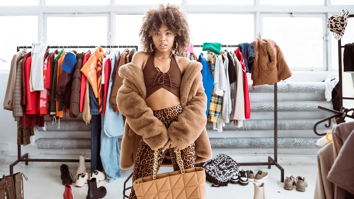 Portrait of confident, curly hair, fashionable young woman wearing brown fake fur coat and leopard print pants, holding bag in hands, standing against hanger with clothes, looking at camera. Influencer selling clothes.