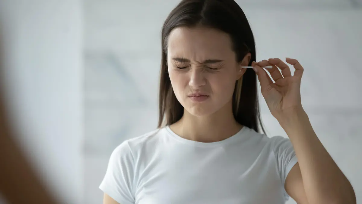 Young woman stand in bathroom look in mirror clean ears with cotton bud, millennial girl feel pain cleansing using stick or swab after shower in home bath, hygiene, daily routine concept