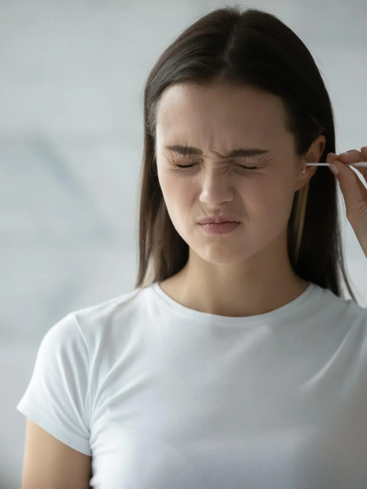 Young woman stand in bathroom look in mirror clean ears with cotton bud, millennial girl feel pain cleansing using stick or swab after shower in home bath, hygiene, daily routine concept